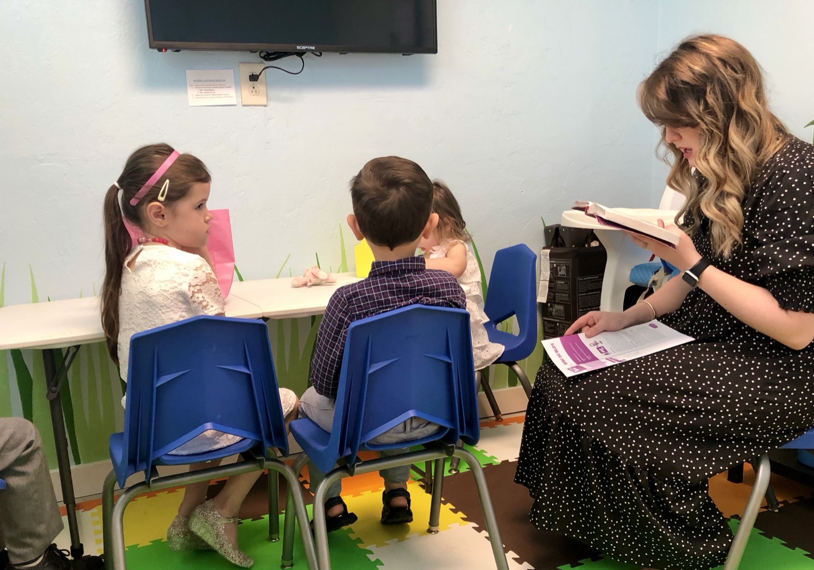 Kendra reading from an open Bible to three young children seated at a classroom table